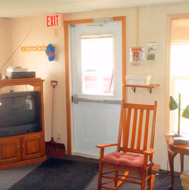 Interior view of a room with a white exit door featuring a window, a wooden rocking chair with a red cushion, a wooden TV stand with an old-style television, a coat rack with a blue cap hanging, a snow shovel leaning against the wall, and a small wooden side table with a green lamp next to a window.