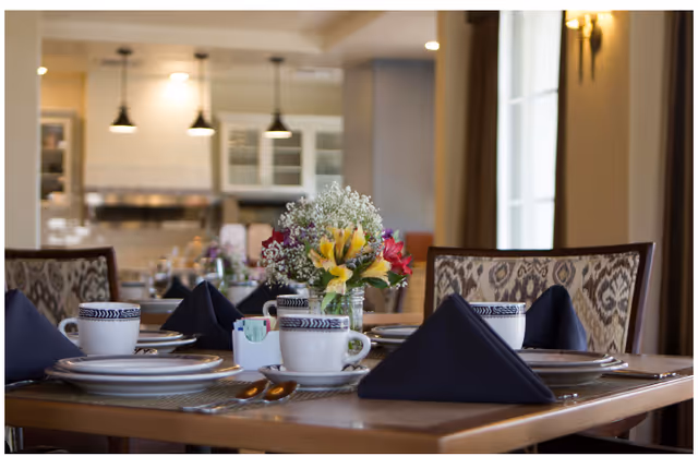 A dining table set with white plates, cups with blue patterns, silverware, and navy blue folded napkins. A small floral centerpiece with yellow, red, and white flowers is placed in the middle of the table. The background shows a kitchen area with pendant lights and cabinets.