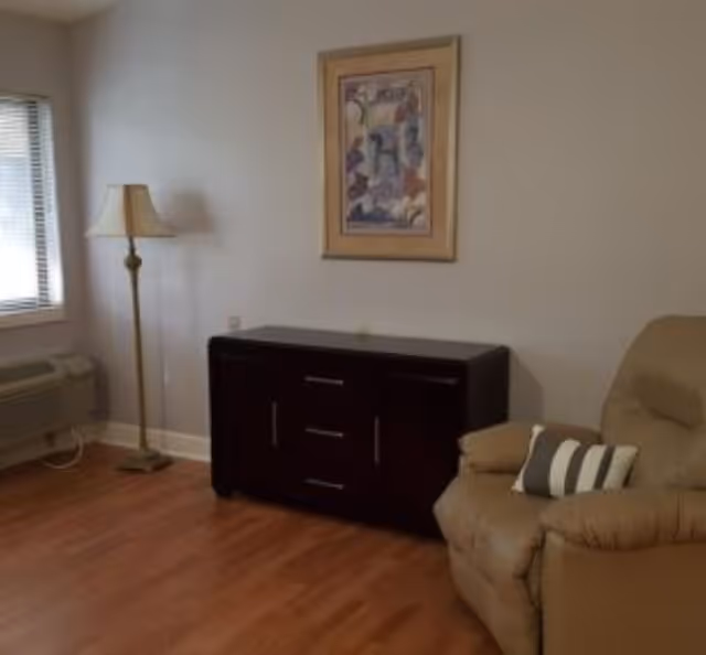 A cozy corner of a living room featuring a beige recliner chair with a striped pillow, a dark wooden cabinet with drawers and doors, a tall floor lamp with a beige lampshade, a window with blinds, and a framed abstract painting on the wall above the cabinet. The floor is wooden.