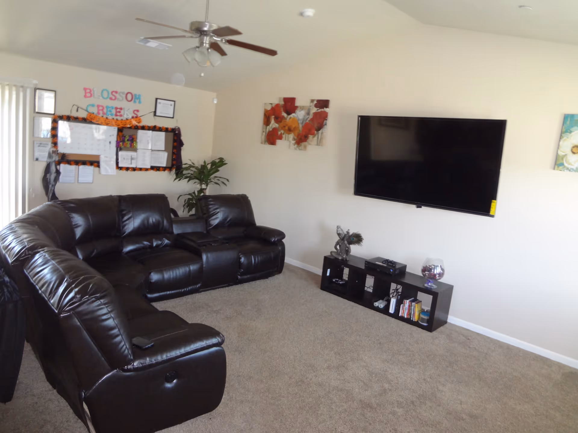 Spacious living room with a black leather sectional, wall-mounted TV, low console, bulletin board and ceiling fan.