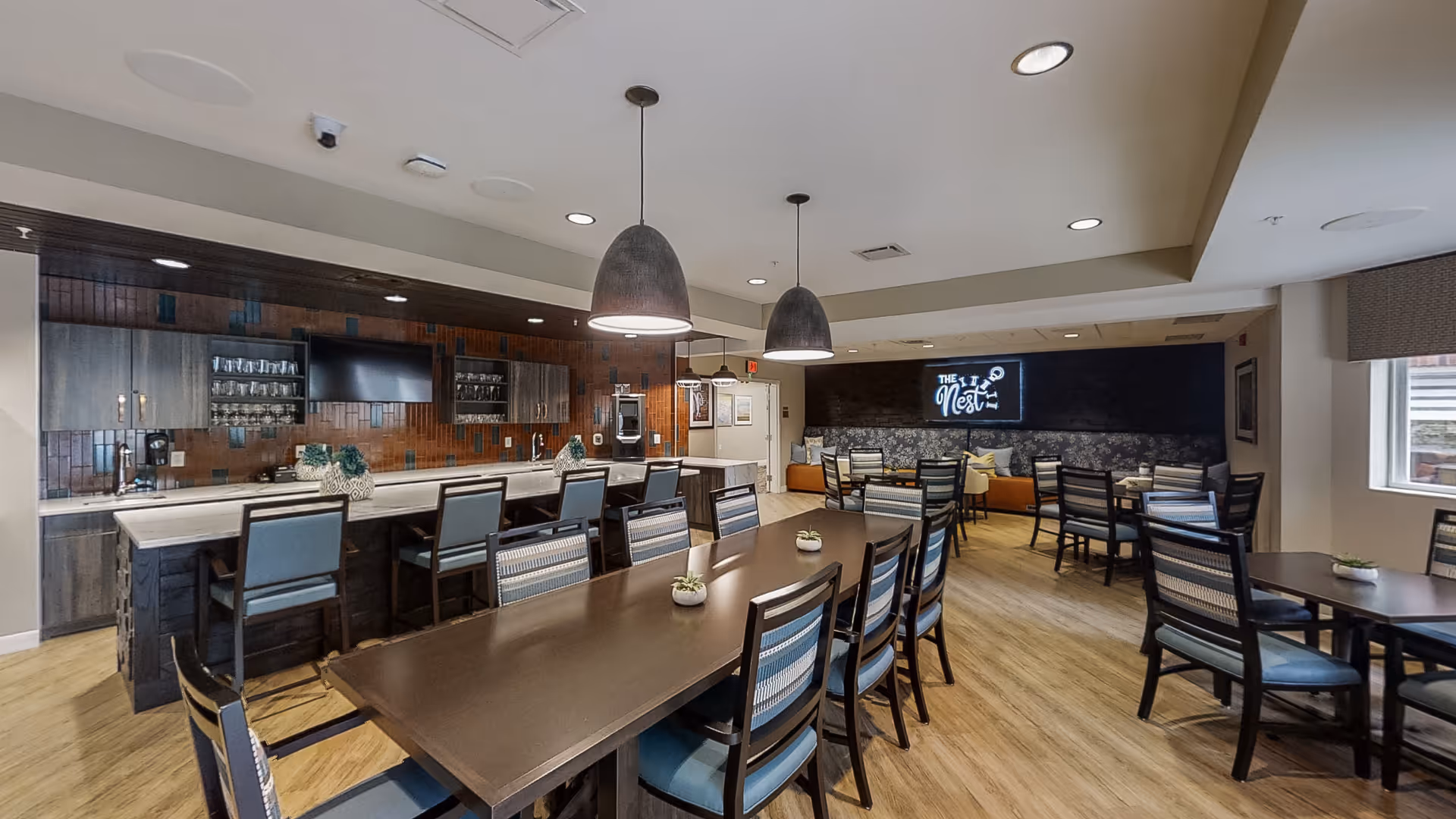 A modern dining area with multiple tables and chairs, a long counter with bar stools, pendant lights hanging from the ceiling, and a wall-mounted TV. The room has wooden flooring and a sign on the back wall that reads 'The Nest'.