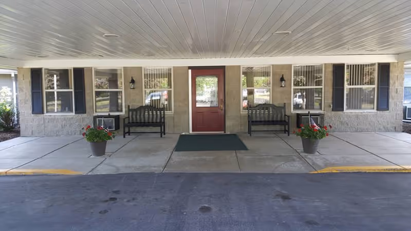 Covered entrance area of a building with a red door in the center, flanked by two black benches and potted plants with red flowers on either side. The building has beige stone walls, multiple windows with black shutters, and a green doormat in front of the door.