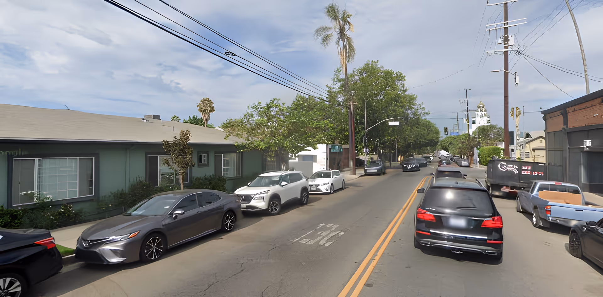 Street view showing parked cars along a road with a single-story green building on the left and various trees and utility poles lining the street under a partly cloudy sky.