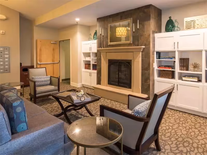 A cozy living room area featuring a stone fireplace with a mantel, flanked by white built-in bookshelves filled with books and decorative items. The room has a patterned carpet, a gray sofa with patterned cushions, two armchairs with cushions, a glass-top side table, and a coffee table with decorative objects. The walls are painted in neutral tones, and there is a wooden door in the background.