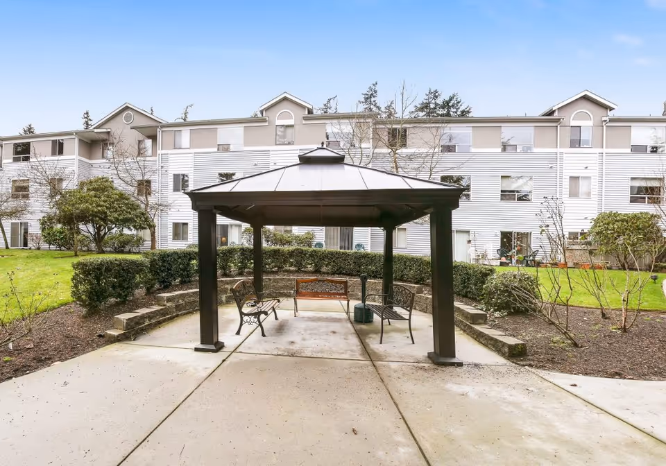 Outdoor seating area with a black metal gazebo covering three benches on a concrete patio, surrounded by trimmed bushes and landscaping. In the background, there is a three-story residential building with white siding and beige accents under a clear blue sky.