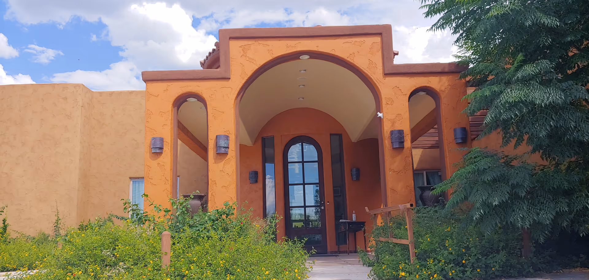 Front entrance of a building with an orange stucco exterior, featuring an arched doorway with glass panels and two smaller arched openings on either side. The entrance is surrounded by green bushes and trees under a partly cloudy blue sky.