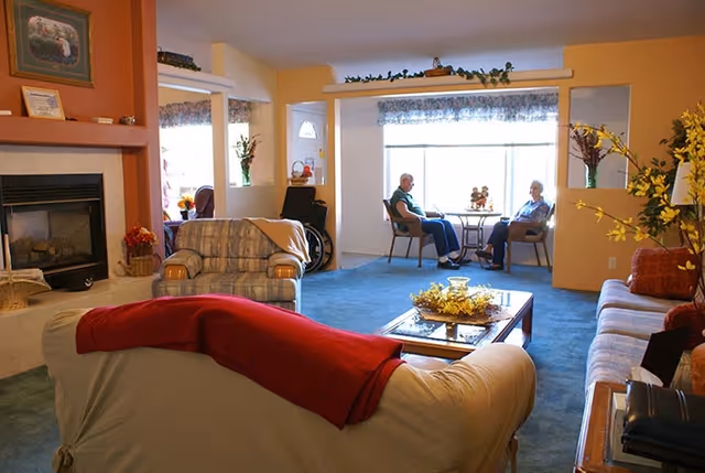 A cozy living room in a senior care facility with multiple sofas and armchairs arranged around a coffee table. Two elderly people are seated at a small table near a large window, engaged in conversation. The room features a fireplace, decorative plants, and warm lighting.