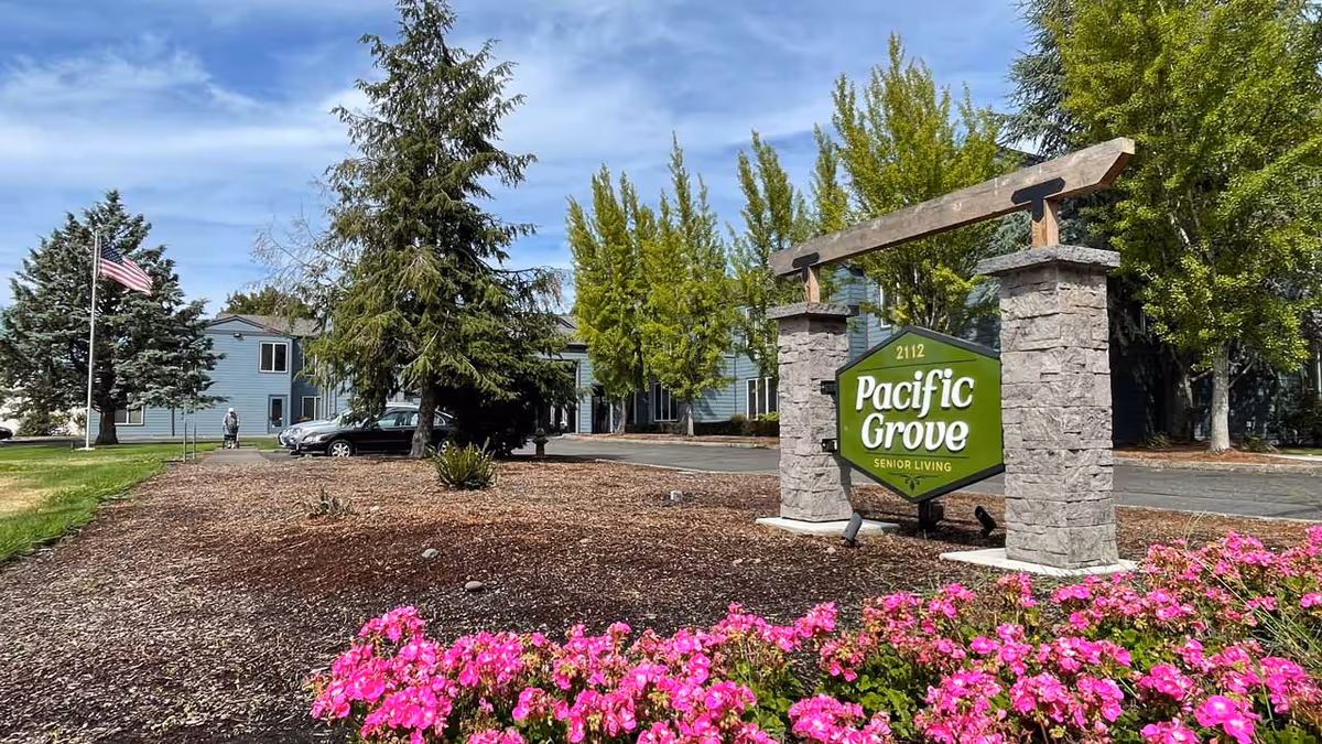 Outdoor view of Pacific Grove Senior Living facility with a green sign mounted on stone pillars, surrounded by pink flowers and trees, with a building and an American flag in the background under a partly cloudy sky.