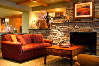 Cozy living room area with a red patterned sofa, wooden coffee table, and a stone fireplace. The room features warm lighting, a framed landscape painting above the fireplace, and decorative items including a statue and flowers.