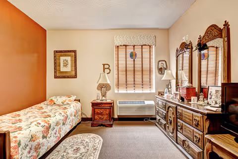 Well-lit furnished bedroom with a single bed, wooden nightstand and dresser, and a window with blinds.