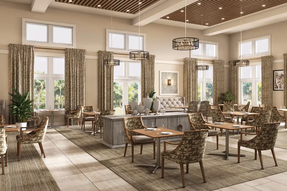 A spacious and well-lit dining room with multiple tables and chairs arranged neatly. The room features large windows with curtains, allowing natural light to fill the space. There are decorative plants and modern pendant lights hanging from a wooden slatted ceiling. The overall decor is warm and inviting with neutral tones.