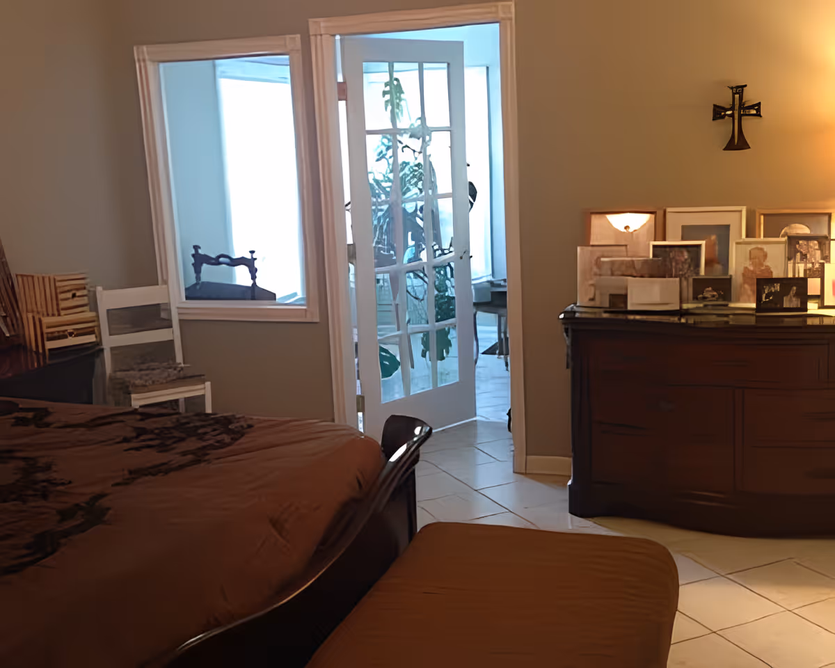 A bedroom with a brown bedspread, a wooden dresser topped with multiple framed pictures, a white chair, and a glass-paneled door leading to a bright room with plants.
