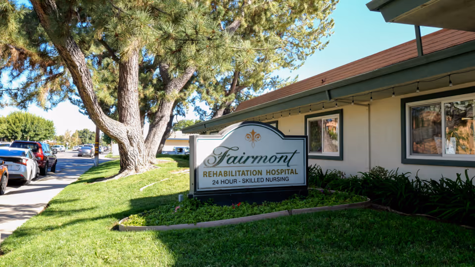 Outdoor view of Fairmont Rehabilitation Hospital sign on a grassy area with trees and parked cars along the street beside the building.