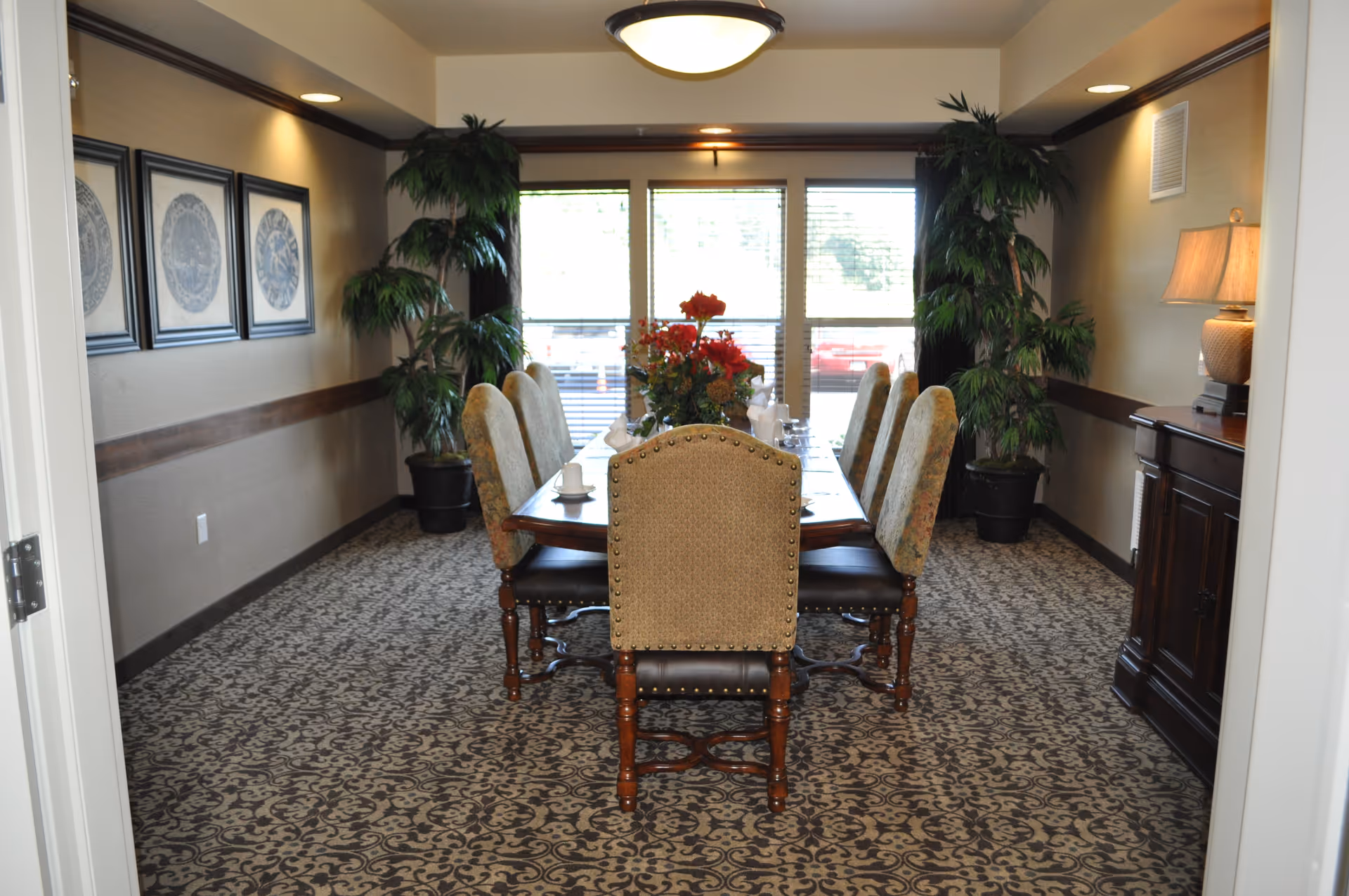A formal dining room with a long wooden table surrounded by eight upholstered chairs. The table is decorated with a floral centerpiece and set with white cups and napkins. The room has patterned carpet, framed artwork on the left wall, two large potted plants near the windows, and a wooden sideboard with a lamp on the right. Large windows with blinds allow natural light to enter the room.