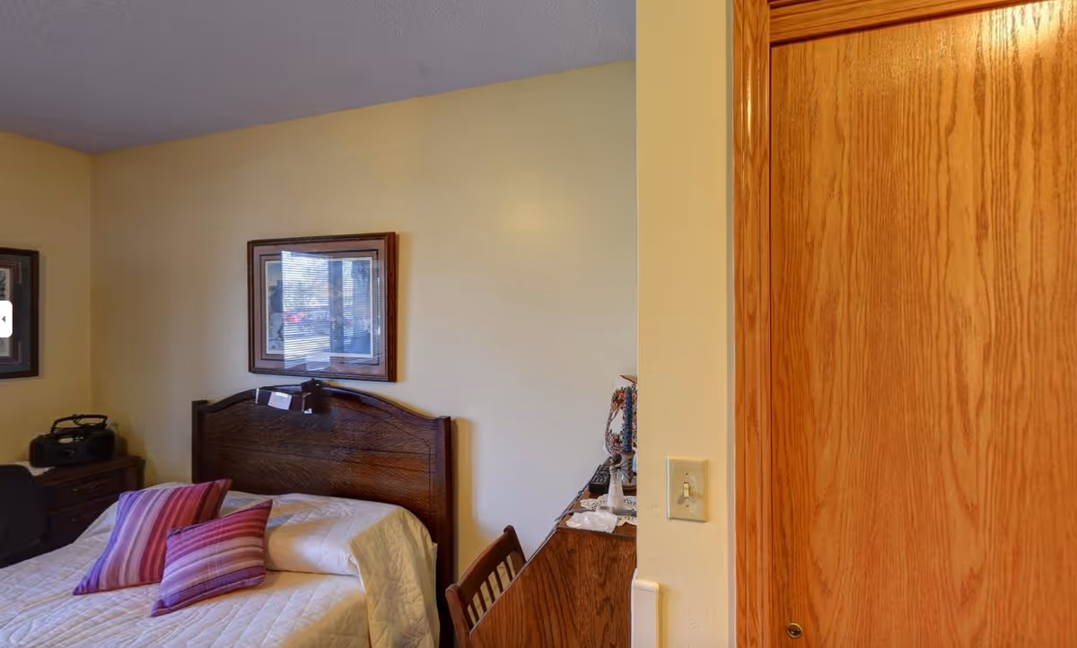 A simple bedroom with a bed against a pale yellow wall, wooden headboard and dresser, framed artwork, and a wooden closet door.