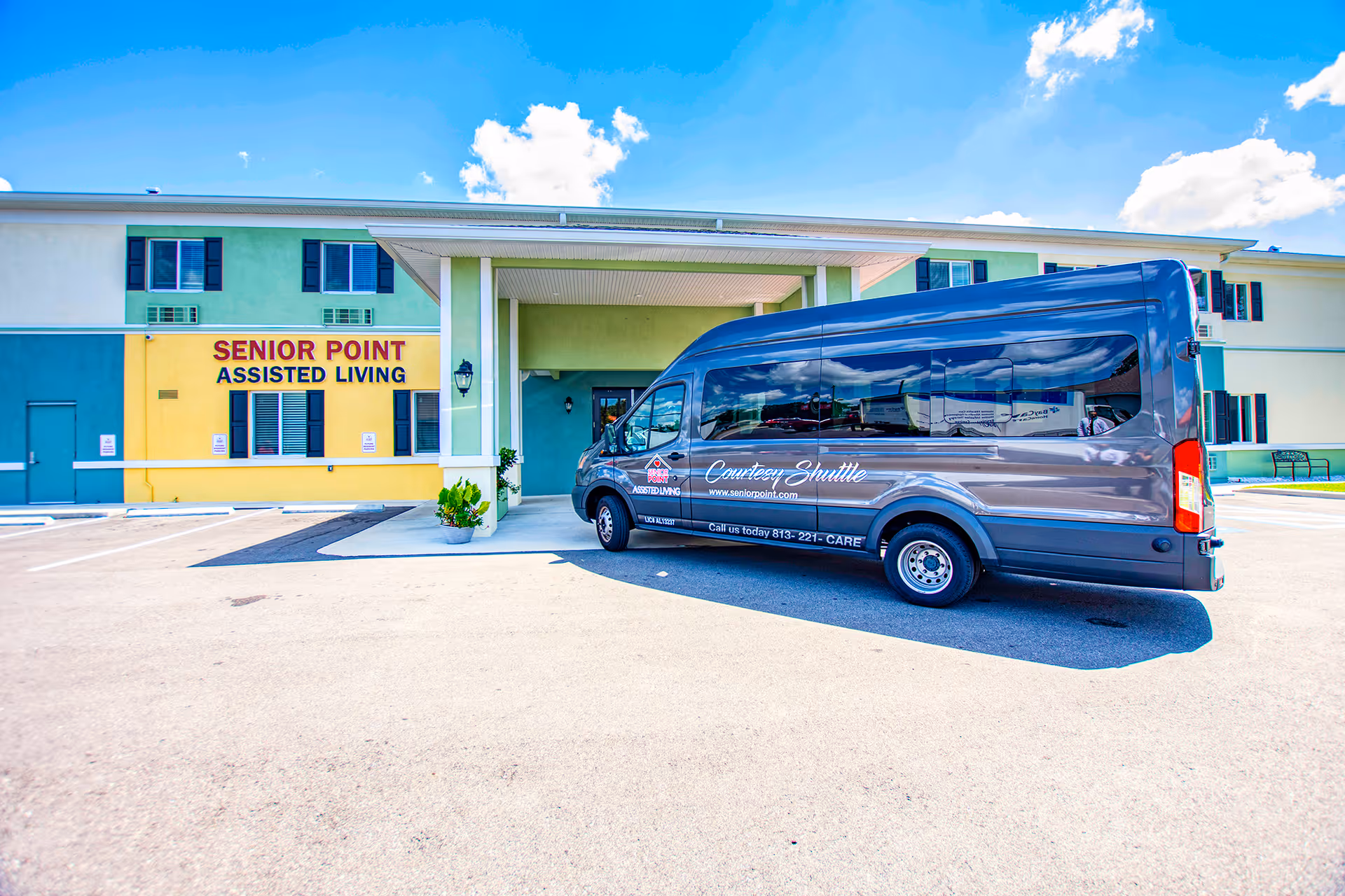 Exterior view of Senior Point Assisted Living facility with a covered entrance and a gray courtesy shuttle van parked in front under a bright blue sky with some clouds.