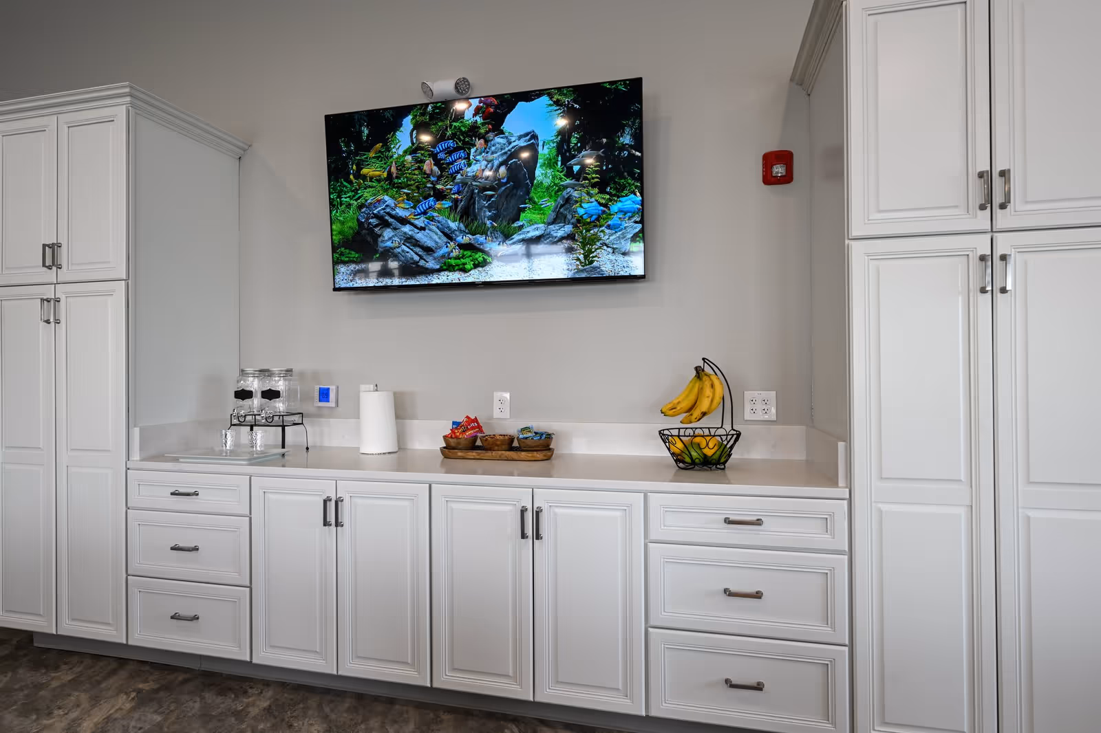 A modern kitchen area with white cabinets and drawers, a countertop holding a paper towel roll, a water dispenser with glasses, a tray with snacks, and a fruit basket with bananas and other fruits. A flat-screen TV mounted on the wall displays an underwater scene with colorful fish and rocks.