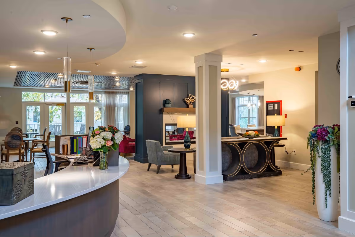 Well-lit senior living facility lobby with a curved reception desk, seating areas, decorative lamps and flowers, and large windows.