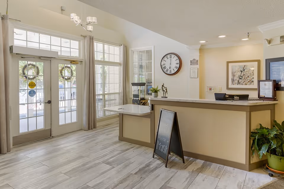 Reception area of a senior living facility with a front desk, a clock on the wall showing 11:50, a small plant on the desk, a framed picture on the wall, large windows with curtains, and double glass doors with wreaths hanging on them.