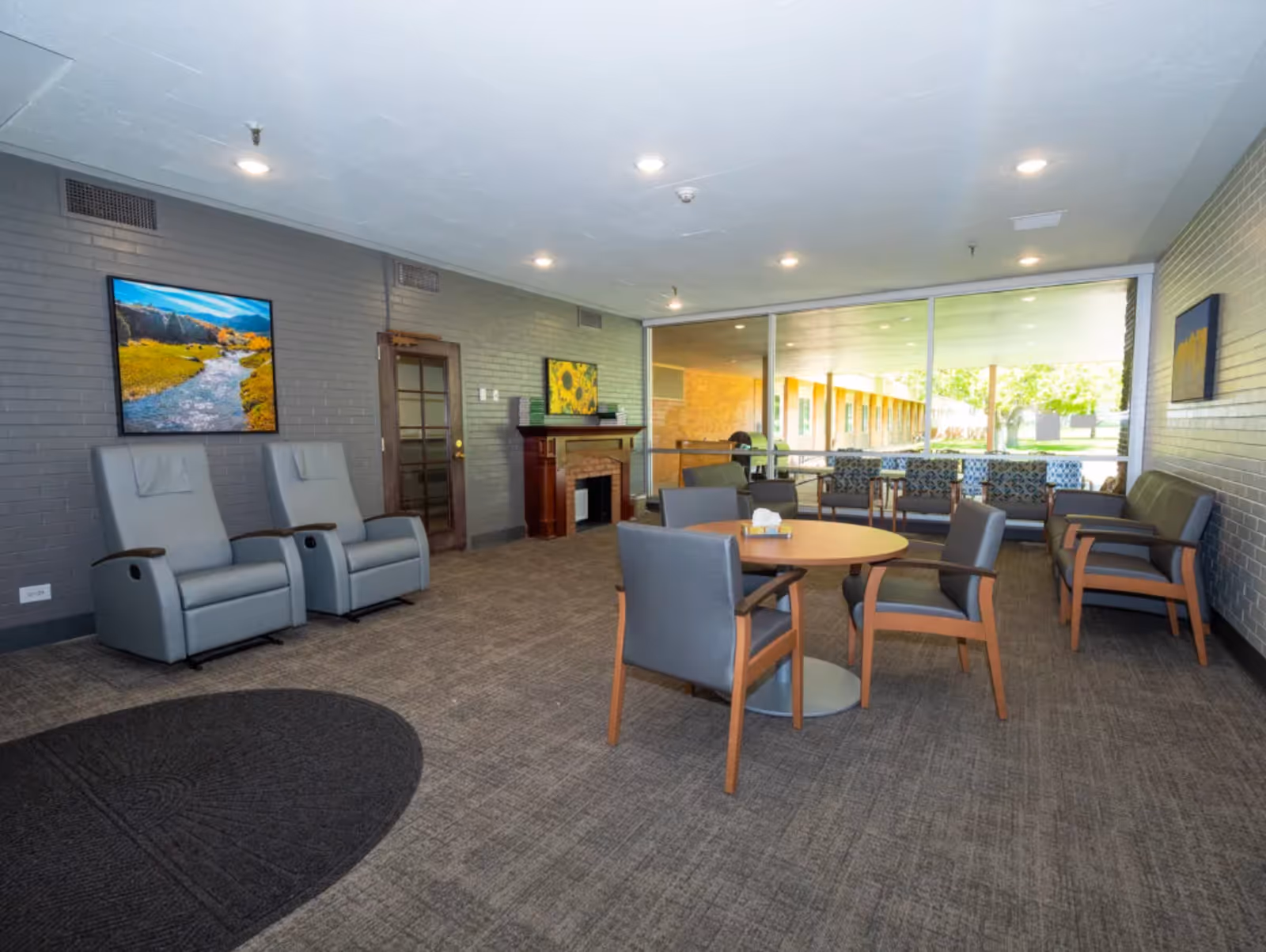 Spacious senior living common lounge with chairs, round table, recliners, a small fireplace and large windows overlooking a courtyard.