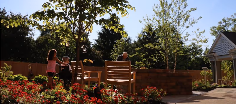 Outdoor garden area with blooming red flowers and green trees. Two elderly people are seated on wooden benches, one in a wheelchair, and a younger woman is standing nearby. The scene is sunlit with a clear blue sky and a building with white latticework is visible on the right.