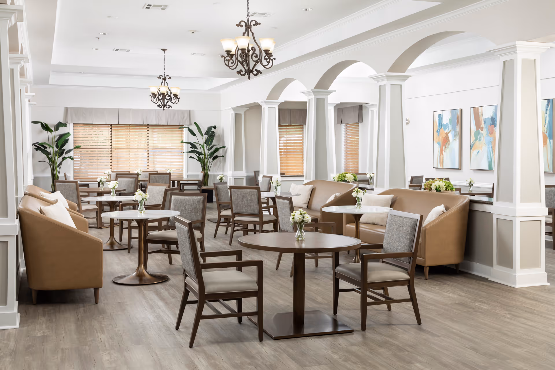 A bright and spacious dining area featuring multiple round wooden tables with gray cushioned chairs and tan leather sofas. The room has large windows with wooden blinds, white walls with decorative molding, and several abstract paintings. Elegant chandeliers hang from the ceiling, and green plants add a touch of nature to the space.