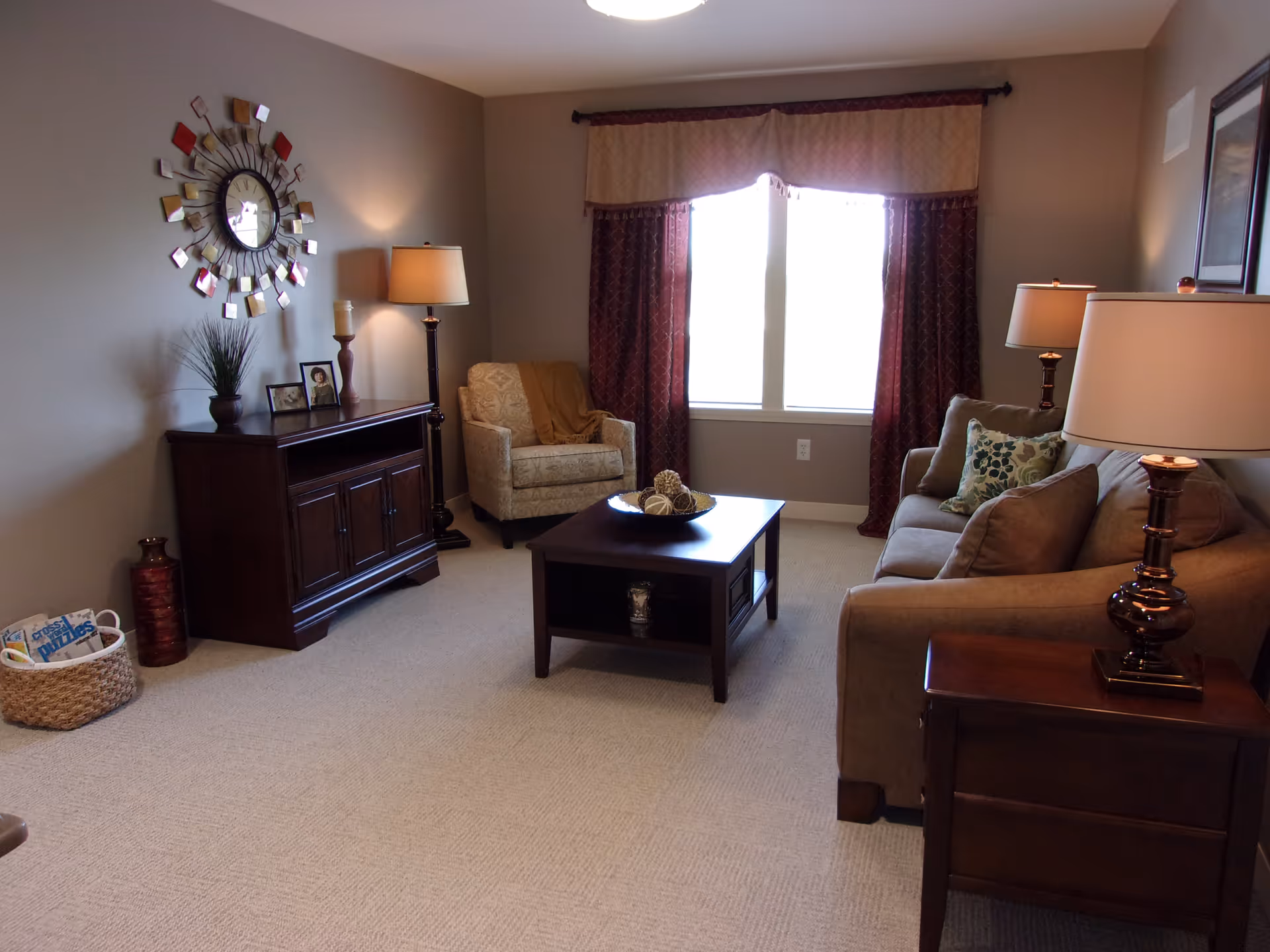 A cozy living room with a beige armchair and a brown sofa adorned with patterned cushions. There is a dark wooden coffee table in the center with decorative items on it. A wooden side table with a lamp is next to the sofa, and another lamp stands beside the armchair. A wooden cabinet with framed photos, a plant, and a candle is against the wall, which also features a decorative wall clock. The window is covered with maroon curtains and a beige valance. A basket with magazines is on the floor near the cabinet.