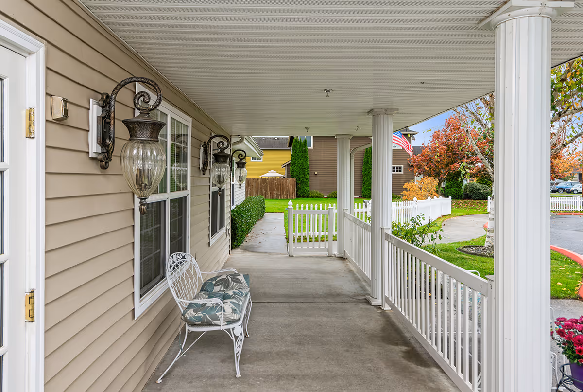 Covered porch of a senior living facility with white railings, chairs, and lamps.
