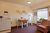 Small senior living suite interior with a round dining table and two chairs, an upholstered armchair, cabinet, floor lamp, window with plants, and framed wall art.