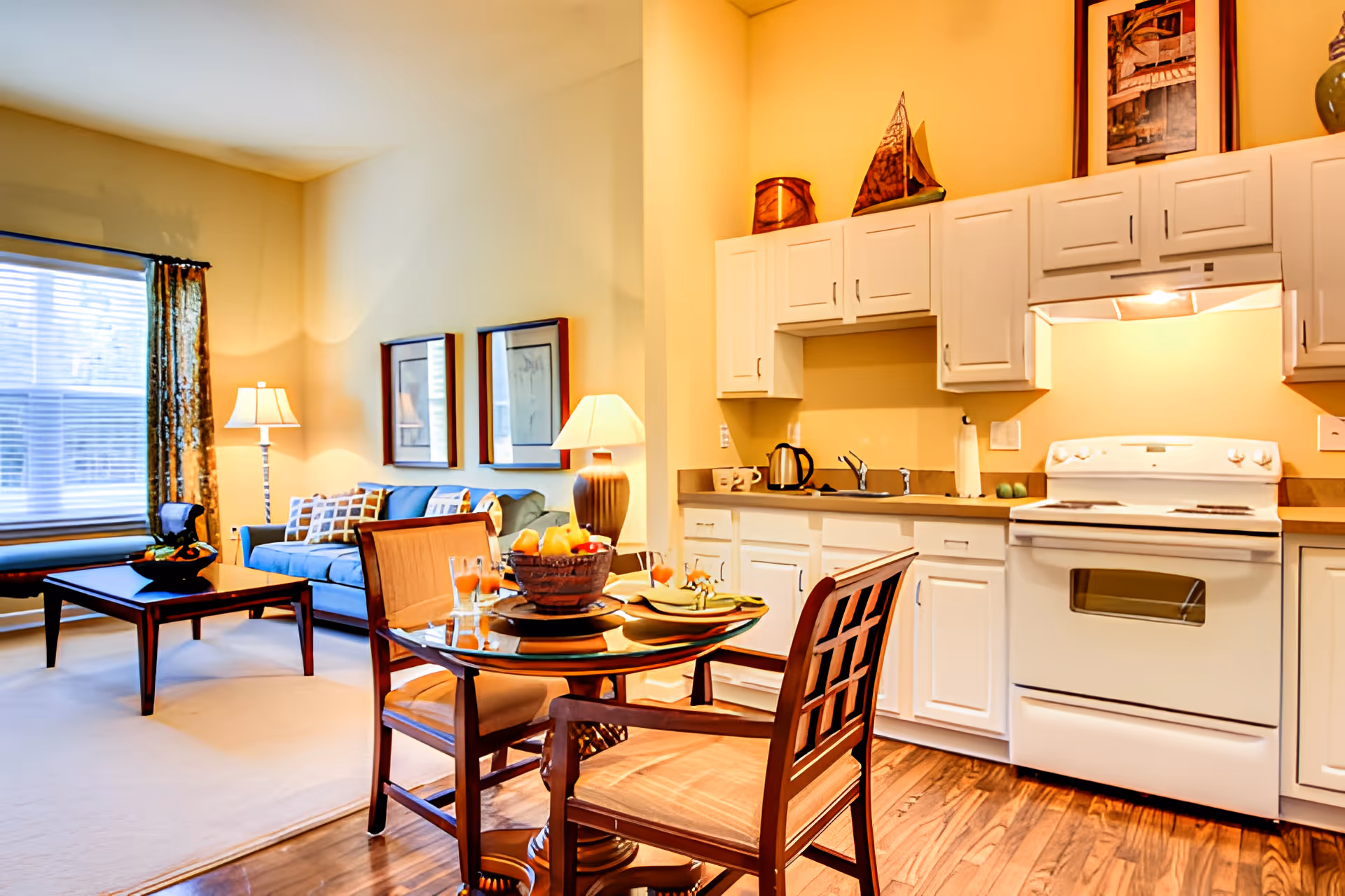 A cozy living space featuring a small kitchen with white cabinets and an electric stove, a round glass dining table with two wooden chairs, and a living room area with a blue sofa, coffee table, floor lamp, and two framed pictures on the wall. A window with curtains allows natural light into the room.