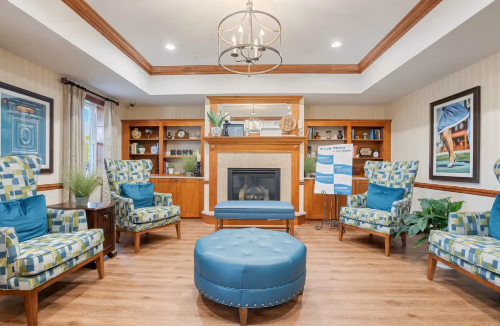 A cozy living room area in a senior living facility with patterned armchairs featuring blue and green squares, blue cushions, a round blue ottoman, and a blue bench in front of a fireplace with wooden built-in shelves on either side. The shelves hold decorative items and plants. There is a chandelier hanging from the ceiling, a framed photo of a person in a blue dress on the wall, and a sign on a stand that reads 'live more at the hearth'. The room has wood flooring and light-colored walls with wooden trim.