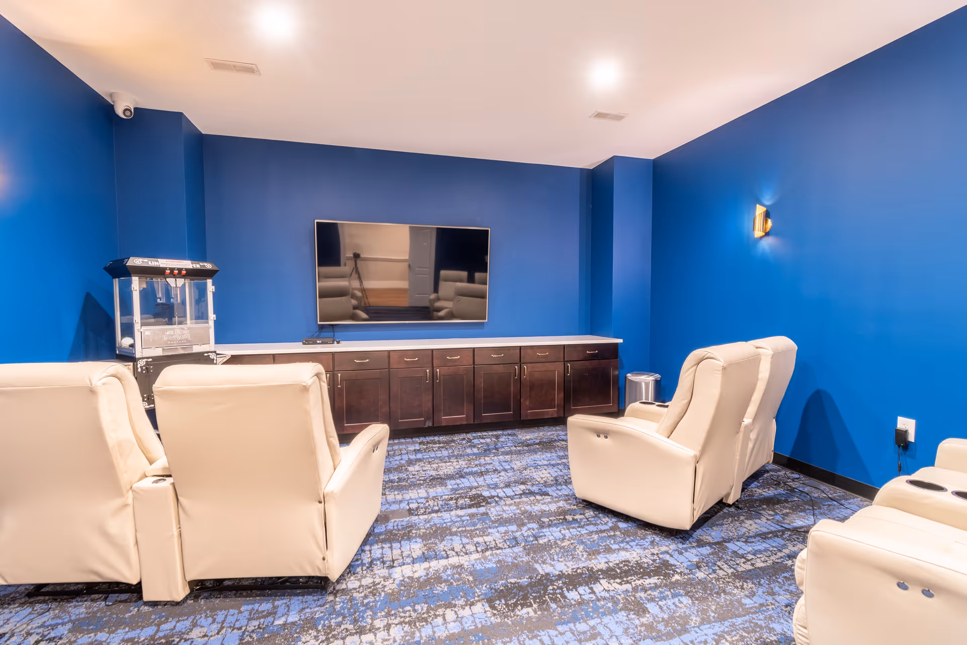 A cozy media room with blue walls and patterned blue carpet, featuring four beige recliner chairs arranged in two rows facing a large flat-screen TV mounted on the wall above a dark wooden cabinet. A popcorn machine is positioned on the left side of the cabinet.