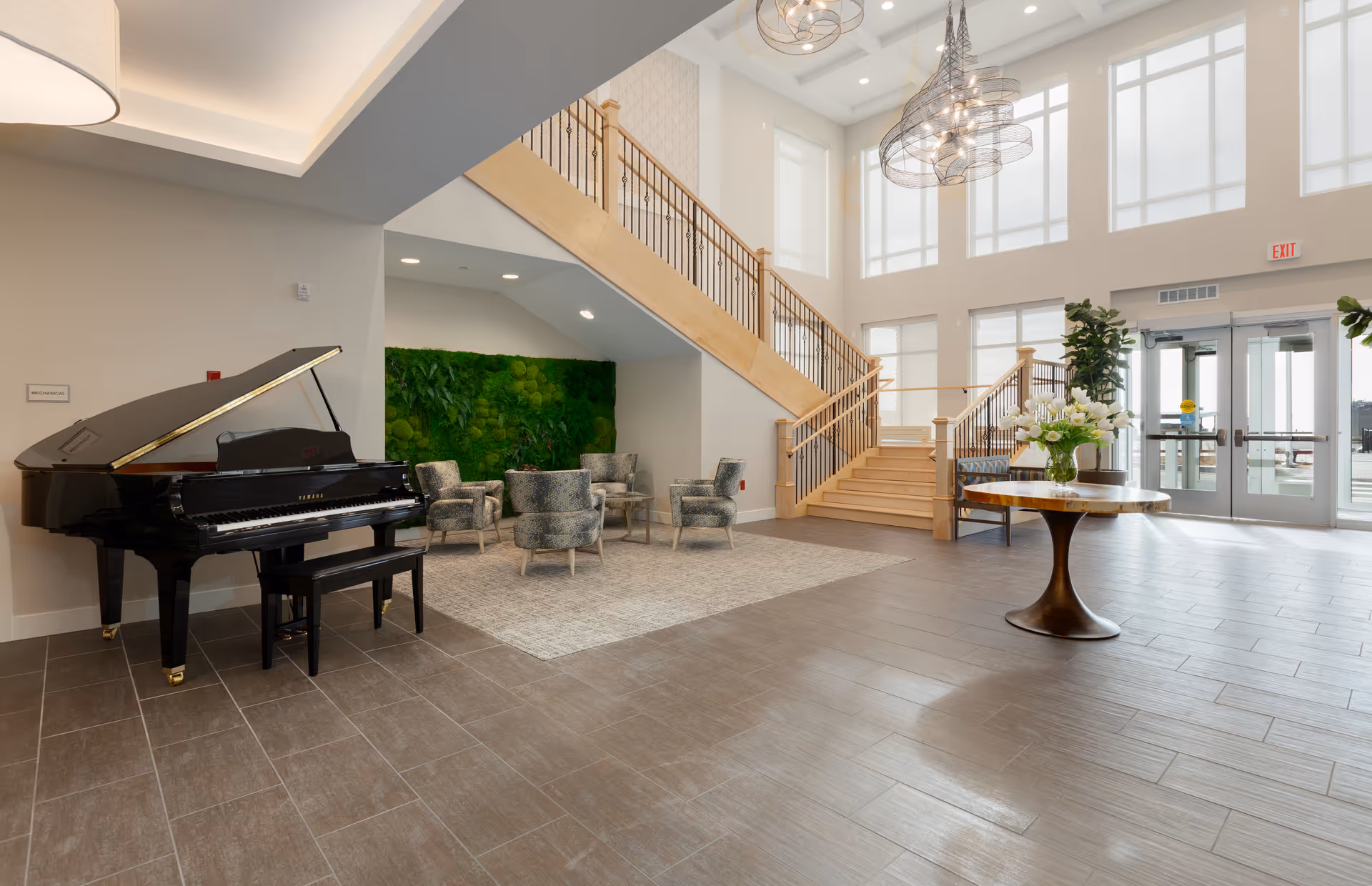 Spacious and bright lobby area with large windows, a grand piano on the left, a seating area with four upholstered chairs and a green moss wall behind them, a wooden staircase with metal railings leading to an upper floor, and a round table with a flower arrangement in the center. The floor is tiled, and modern pendant lights hang from the ceiling.