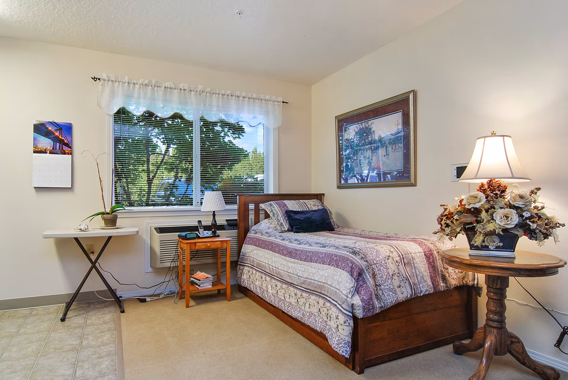 A neatly furnished bedroom with a single bed, bedside tables, a lamp, and a window showing trees outside.