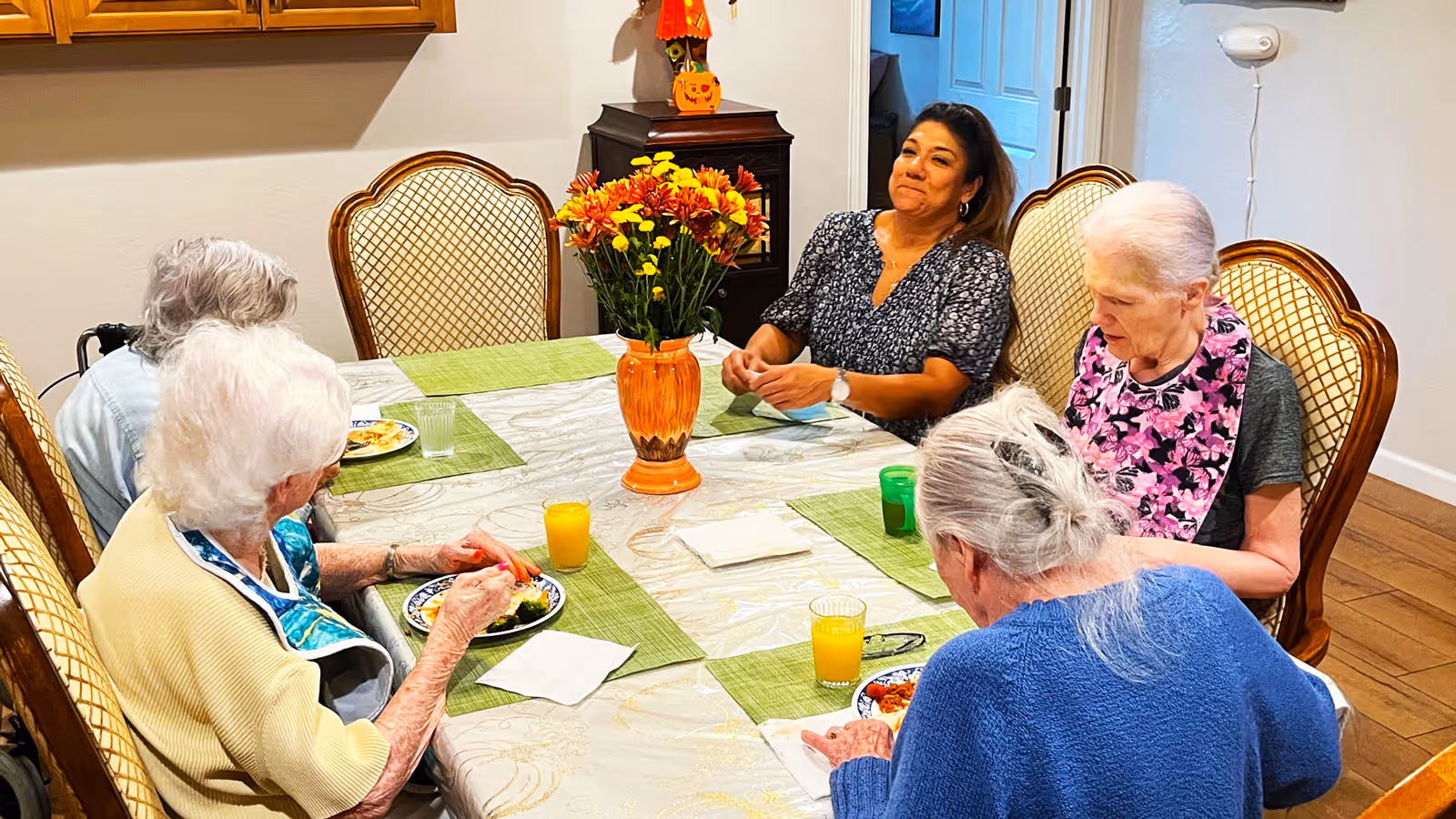 Four elderly women and one caregiver sitting around a dining table with green placemats and plates of food. A vase with orange and yellow flowers is in the center of the table. The setting appears warm and home-like with wooden chairs and a cabinet in the background.