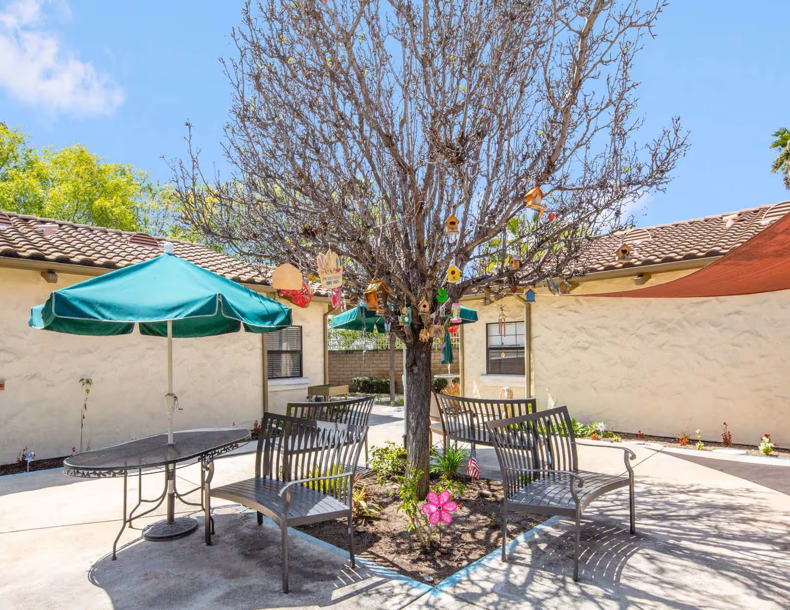 Outdoor courtyard area with a leafless tree decorated with multiple colorful birdhouses and ornaments. Surrounding the tree are metal benches and a table with a green umbrella. The courtyard is enclosed by beige stucco walls with tiled roofs under a clear blue sky.