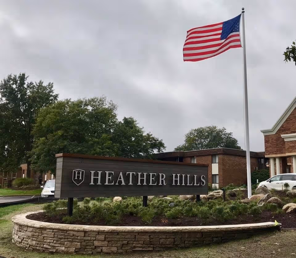 Outdoor view of The Village of Heather Hills facility sign with an American flag on a flagpole and a brick building in the background under a cloudy sky.
