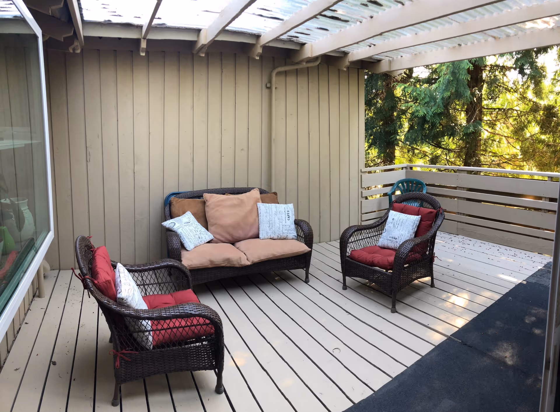 Covered outdoor patio area with beige wooden flooring and walls, featuring a wicker loveseat and two wicker chairs with red and beige cushions. The patio is surrounded by a railing and overlooks green trees.