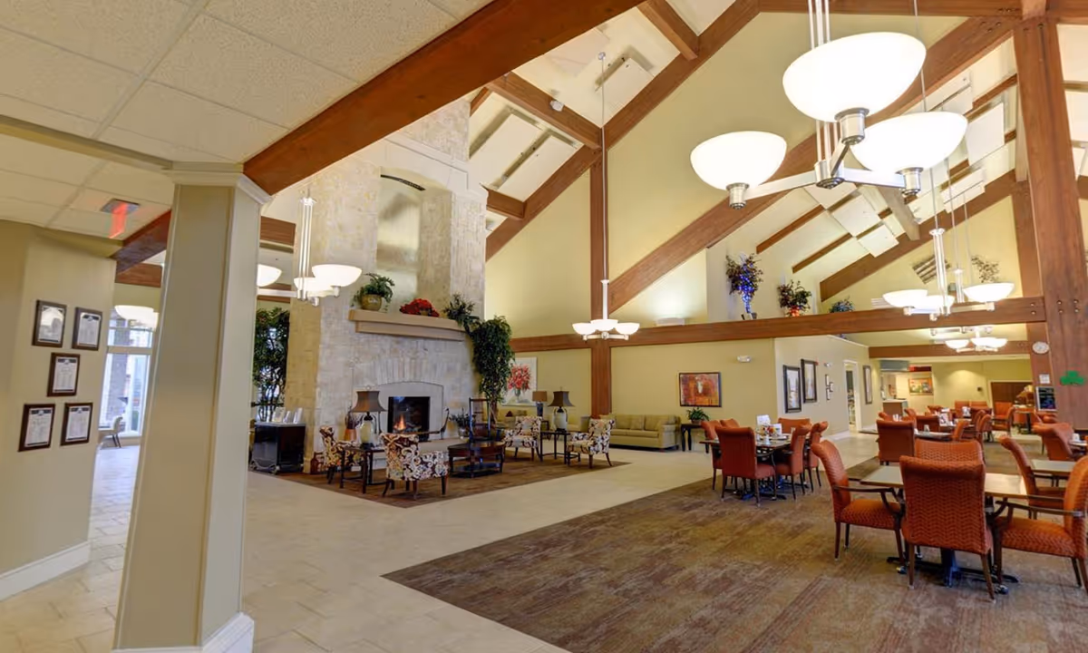Spacious senior living facility common area with high vaulted ceilings featuring exposed wooden beams, multiple hanging light fixtures, a large stone fireplace with a fire burning, comfortable seating including patterned armchairs and sofas, and several dining tables with orange upholstered chairs.