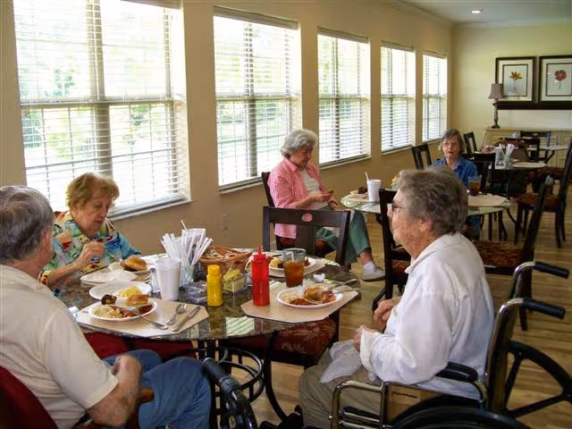 A group of elderly people sitting around tables in a well-lit dining area with large windows. They are eating meals and drinking beverages. Some individuals are seated in wheelchairs. The room has wooden flooring and framed artwork on the walls.