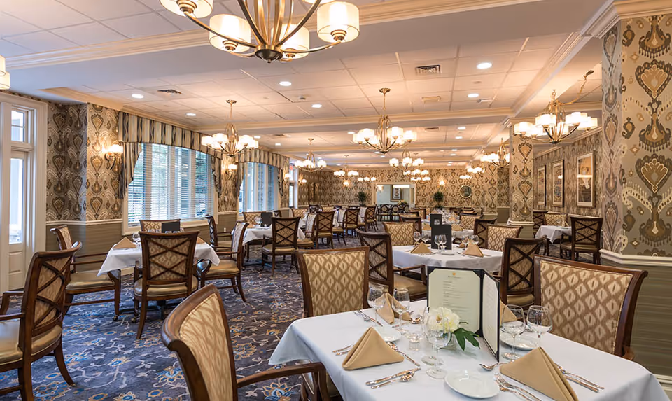 Elegant dining room with multiple tables covered in white tablecloths, set with beige napkins, glassware, and menus. The room features patterned wallpaper, large windows with blinds, ornate chandeliers, and upholstered wooden chairs arranged neatly around the tables.