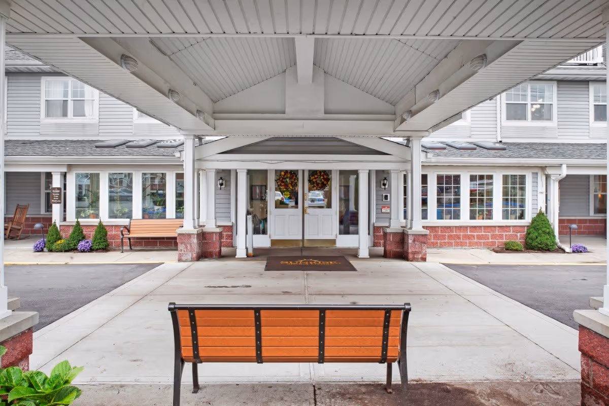 Covered front entrance with a bench facing double doors decorated with wreaths at a senior living facility.