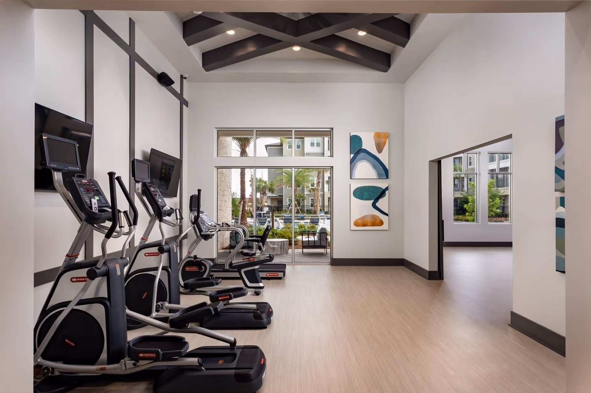 A modern fitness room with several elliptical machines lined up against a wall with mounted flat-screen TVs. The room has light wood flooring, white walls with dark trim, and a high ceiling with exposed beams. Large windows and glass doors at the far end provide a view of an outdoor area with palm trees and seating.