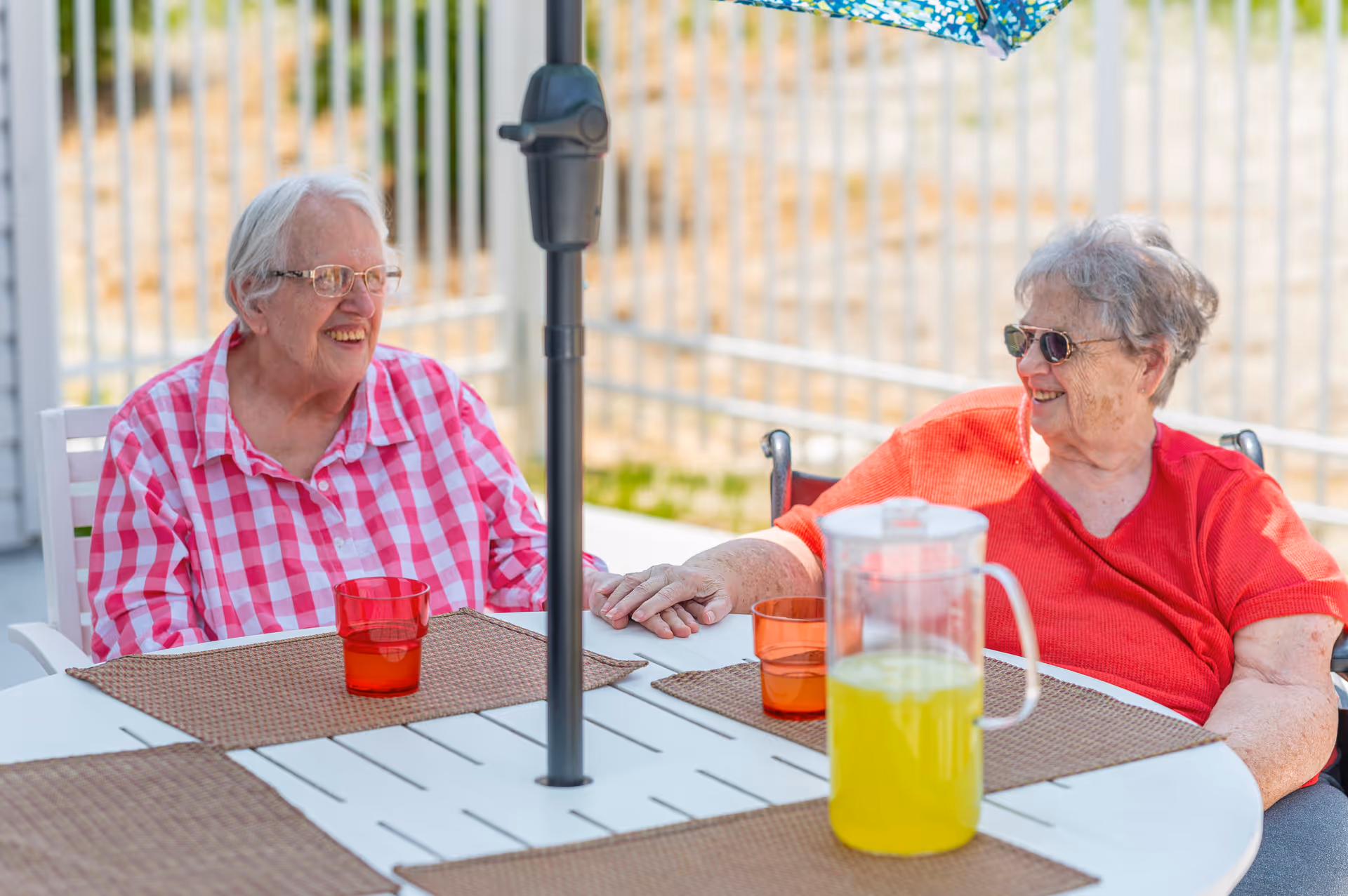 Two elderly women sitting outdoors at a round table with placemats, holding hands and smiling at each other. One woman is wearing a red and white checkered shirt and glasses, while the other is wearing a red shirt and sunglasses. There is a pitcher of yellow drink and two orange cups on the table, with a colorful umbrella providing shade.