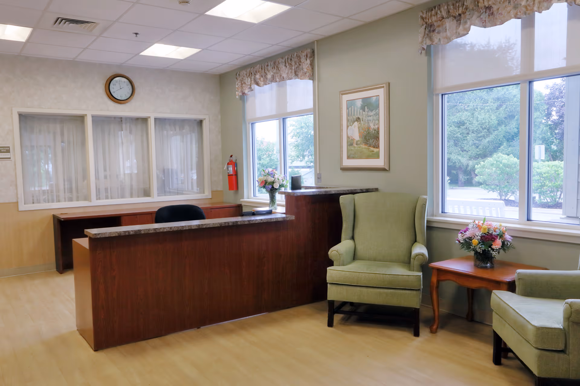 Reception area with a wooden desk and a black office chair behind it. Two green upholstered armchairs are positioned near a window with floral curtains. A small wooden table between the chairs holds a vase of flowers. A clock is mounted on the wall above a set of windows with white curtains. A fire extinguisher is mounted on the wall near the window.