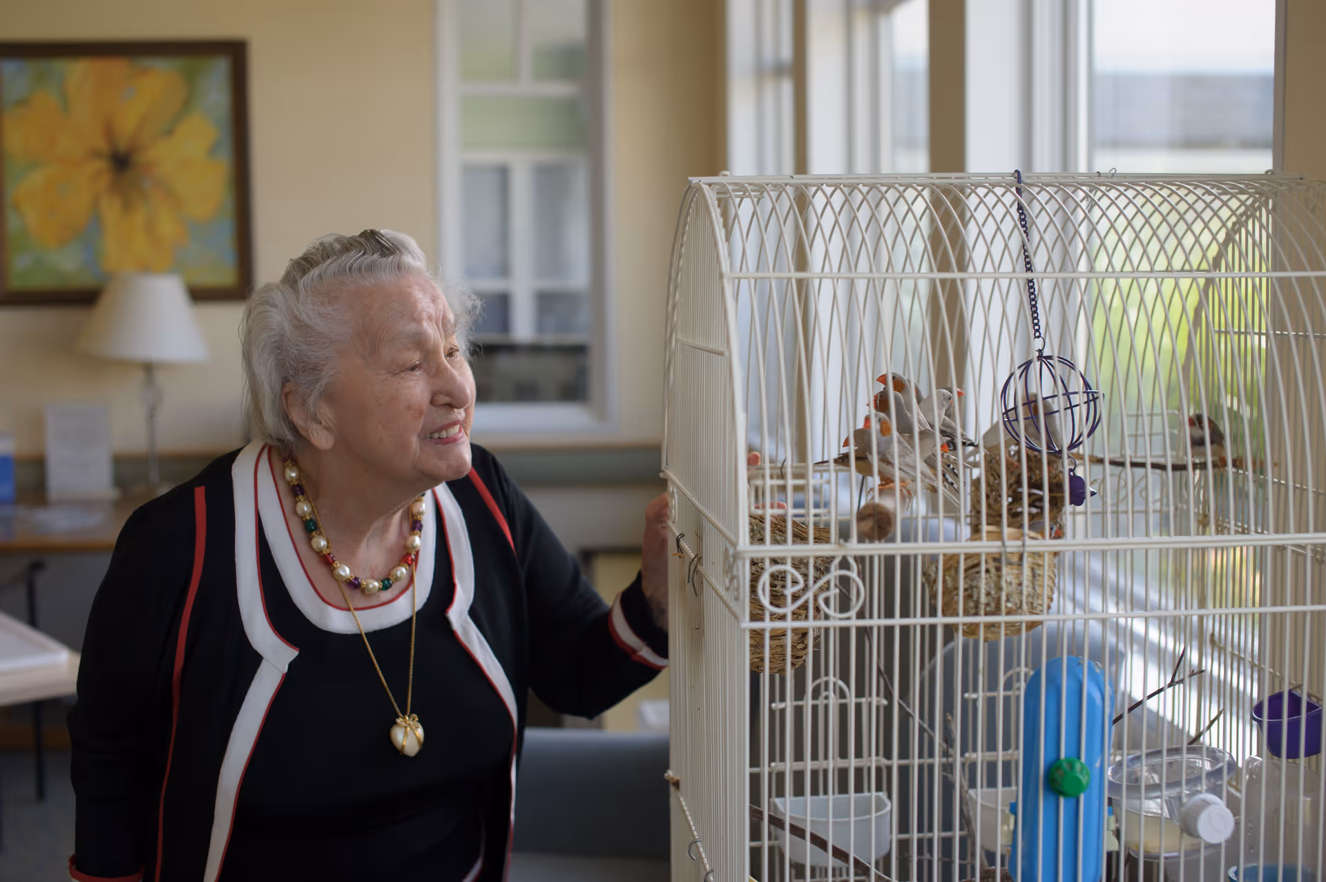 An elderly woman with gray hair and a colorful beaded necklace is smiling and looking at a white birdcage containing several small birds inside a well-lit room with large windows and a painting on the wall.