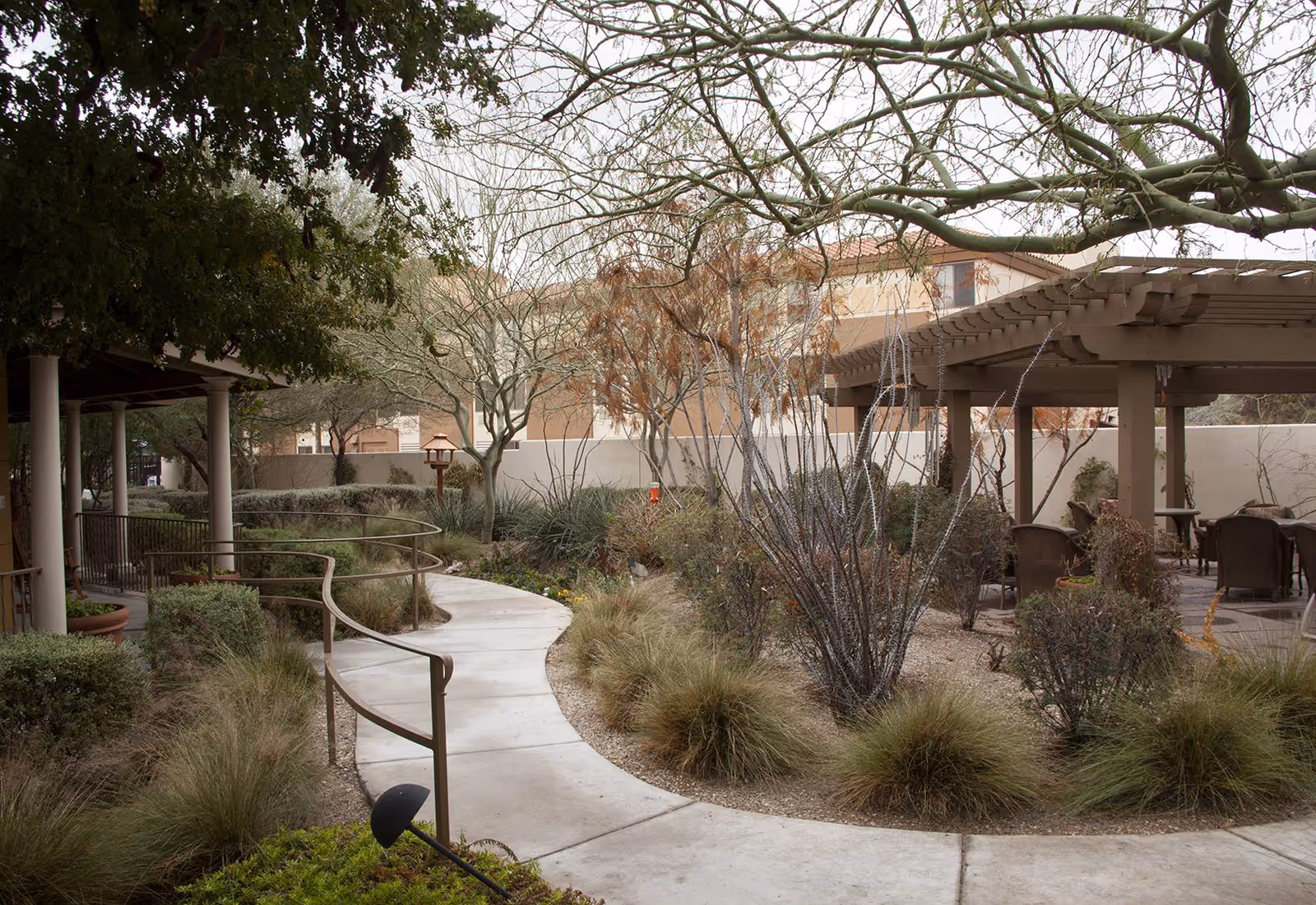 A winding concrete pathway with metal handrails curves through a landscaped outdoor garden area with various shrubs, grasses, and leafless trees. To the right, there is a wooden pergola with several chairs and tables underneath. The sky is overcast, and a building is visible in the background.