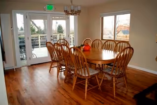 A dining room with a large wooden oval table surrounded by ten wooden chairs with cushions. There is a chandelier hanging above the table and a decorative centerpiece on the table. The room has wooden flooring and large glass doors leading to an outdoor patio area with a railing and trees visible outside.