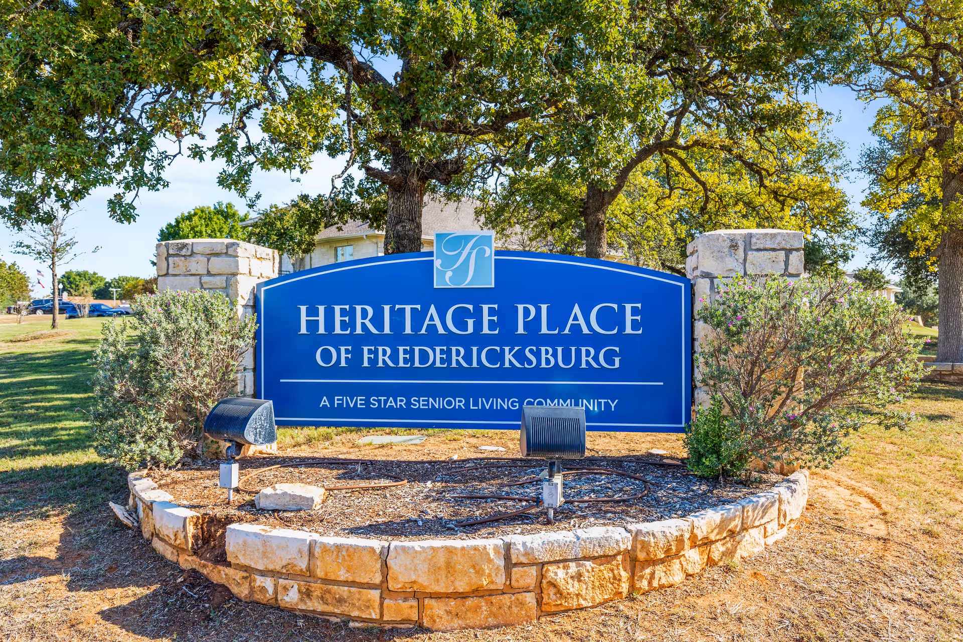 A large blue sign for Heritage Place of Fredericksburg, a five star senior living community, set in a landscaped area with stone edging, bushes, and trees in the background under a clear sky.