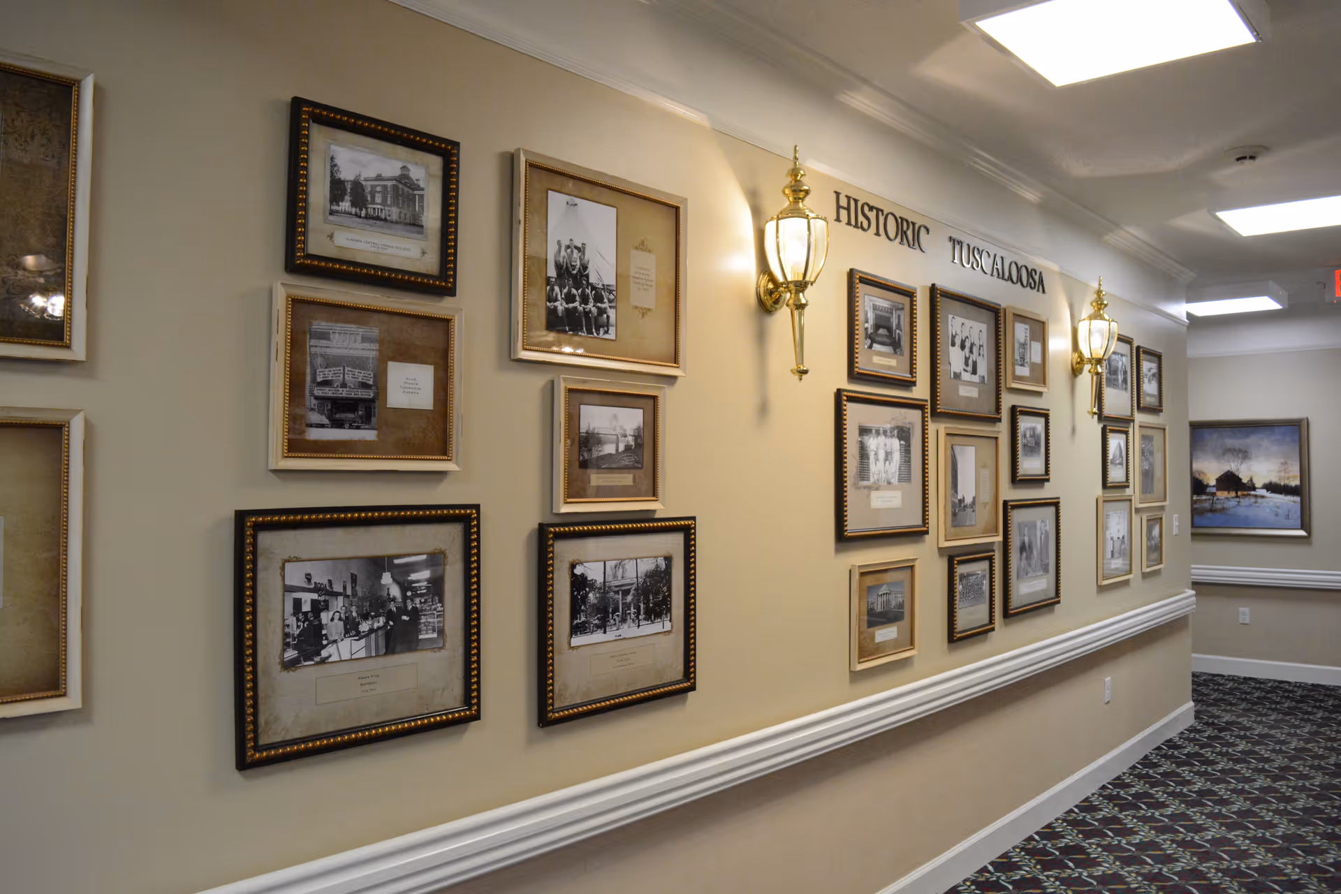 A hallway in a senior living facility with framed historical black and white photographs on the wall under the heading 'Historic Tuscaloosa'. The wall is decorated with two ornate wall lamps, and the floor is carpeted with a patterned design.